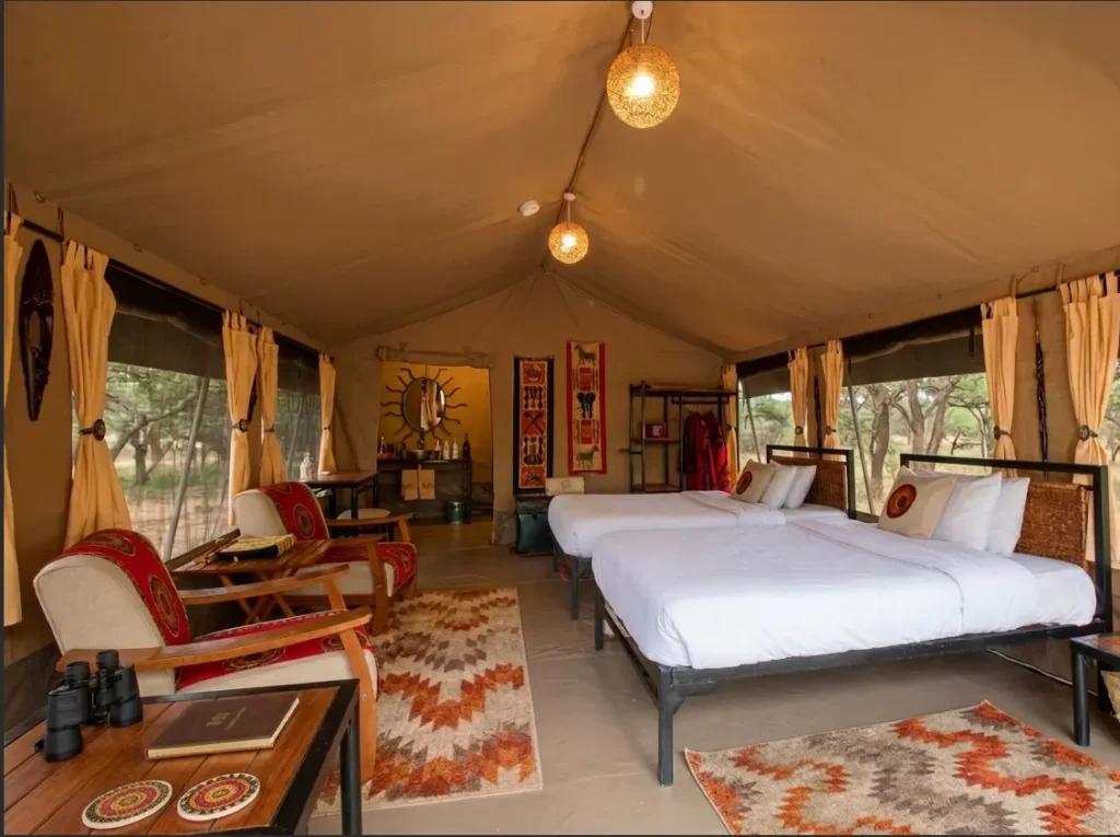 Interior view of an elevated luxury canvas tent at Mara Eden Safari Camp featuring twin beds, native patterned rugs, armchairs, and a rear vanity mirror.